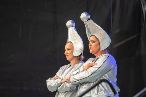 Dwie aktorki w kosmicznych srebrnych strojach z dziwnymi nakryciami na głowie stoją z założonymi rękami.